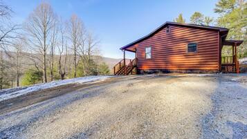 A rustic cabin with a gravel driveway, wooden deck, and a scenic mountain backdrop.