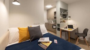 A compact bedroom with a bed, bedside table, and a desk area with a laptop, chair, and a small kitchenette. - Student Only Seren (Swansea)