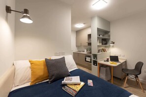 Ein gemütliches Schlafzimmer mit Bett, Nachttisch und einem Arbeitsbereich mit Laptop, Stuhl und einer kleinen Küchenzeile.