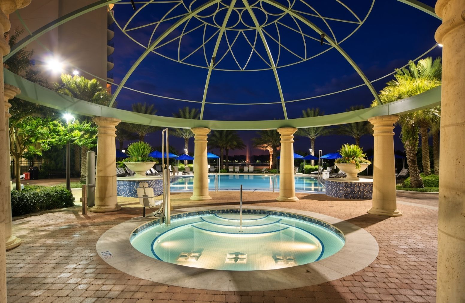 Une piscine circulaire au fond en verre, entourée de palmiers et de colonnes, et recouverte d’une structure en forme de dôme de verre.