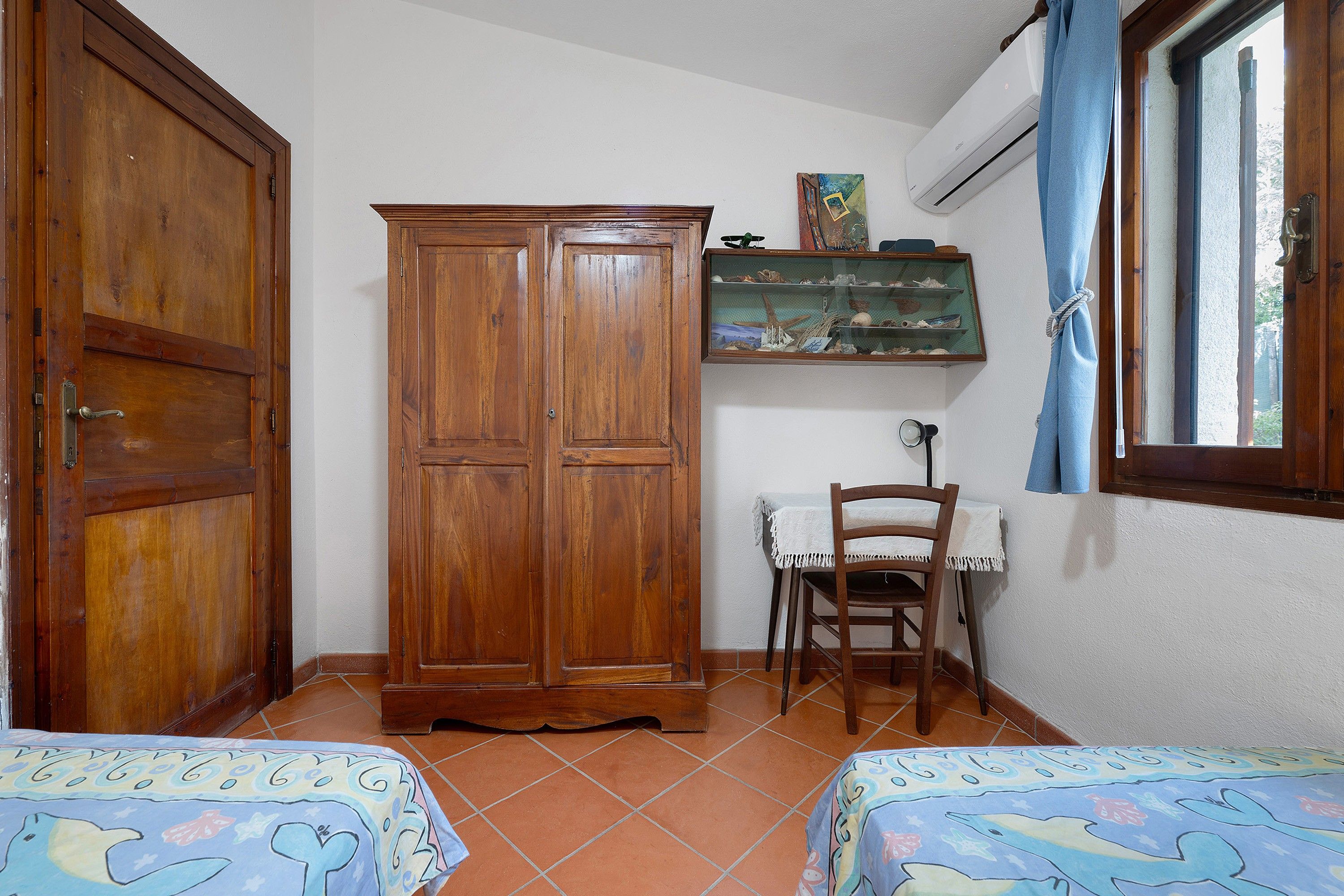Una habitación con dos camas, un armario de madera, una silla, una mesita y una estantería con adornos.