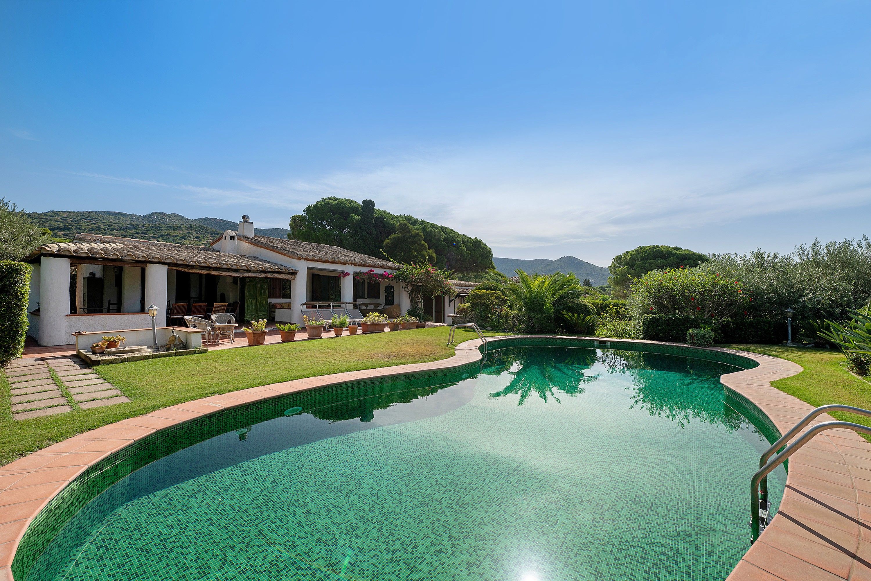 A swimming pool with a tiled edge, a patio with chairs, and a building with a tiled roof.