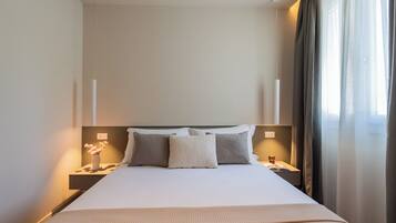 Una habitación de hotel con una cama grande, dos mesitas de noche, un florero con flores y una ventana con cortinas.