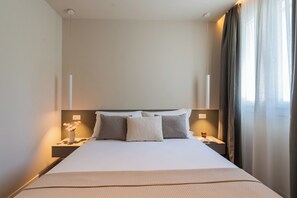 Quarto de hotel com uma cama grande, duas mesas de cabeceira, um criado-mudo, um vaso com flores e uma janela com cortinas.
