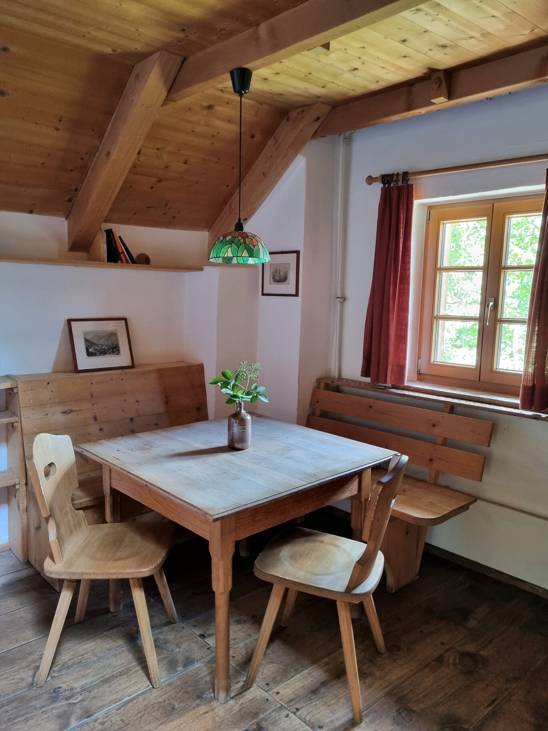 Et spiseområde i træ med et bord og bænke, en hængende lampetilslutning og et vindue med gardiner.