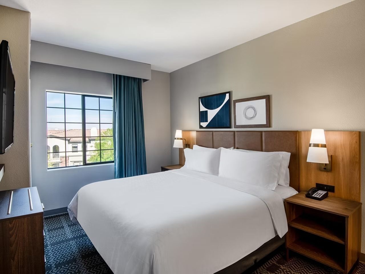 Een hotelkamer met een groot bed, een bureau, een stoel, een raam met gordijnen en een aan de muur bevestigd telefoontoestel.