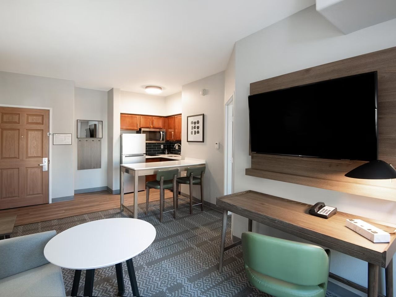 A compact hotel room with a kitchenette, a dining area with a table and chairs, a TV mounted on the wall, and a desk with a lamp.