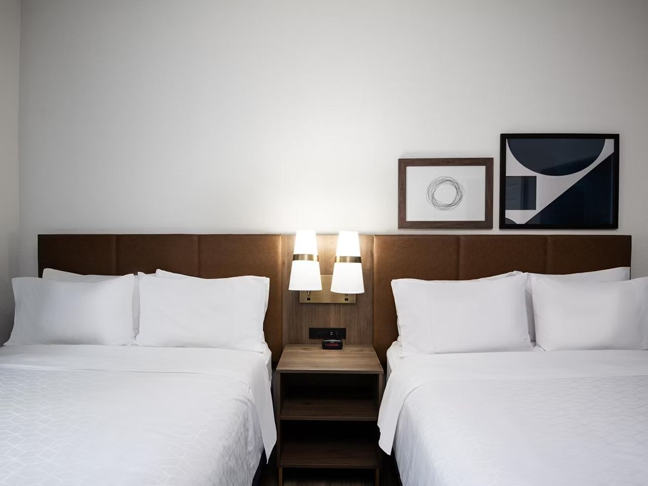 A hotel room with two beds, a nightstand with lamps, a clock, and two framed pictures on the wall.