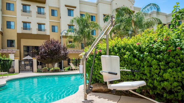 A swimming pool with a sun lounger and a metal chair, surrounded by greenery and a building with balconies.