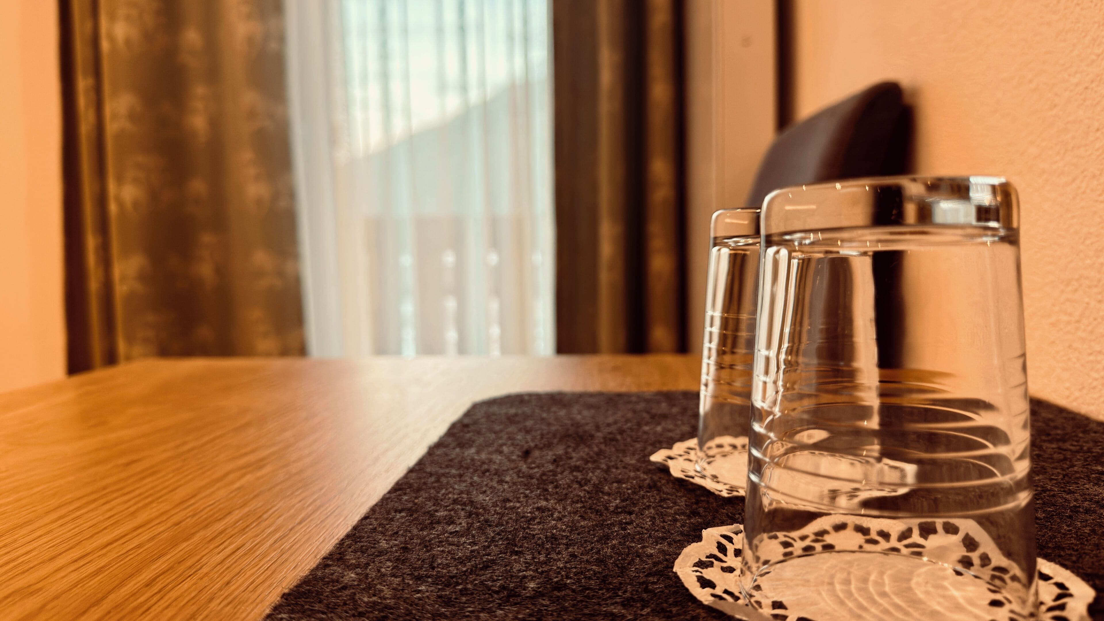 Dos vasos transparentes sobre una mesa de madera con un mantel individual, cerca de una ventana con persianas.