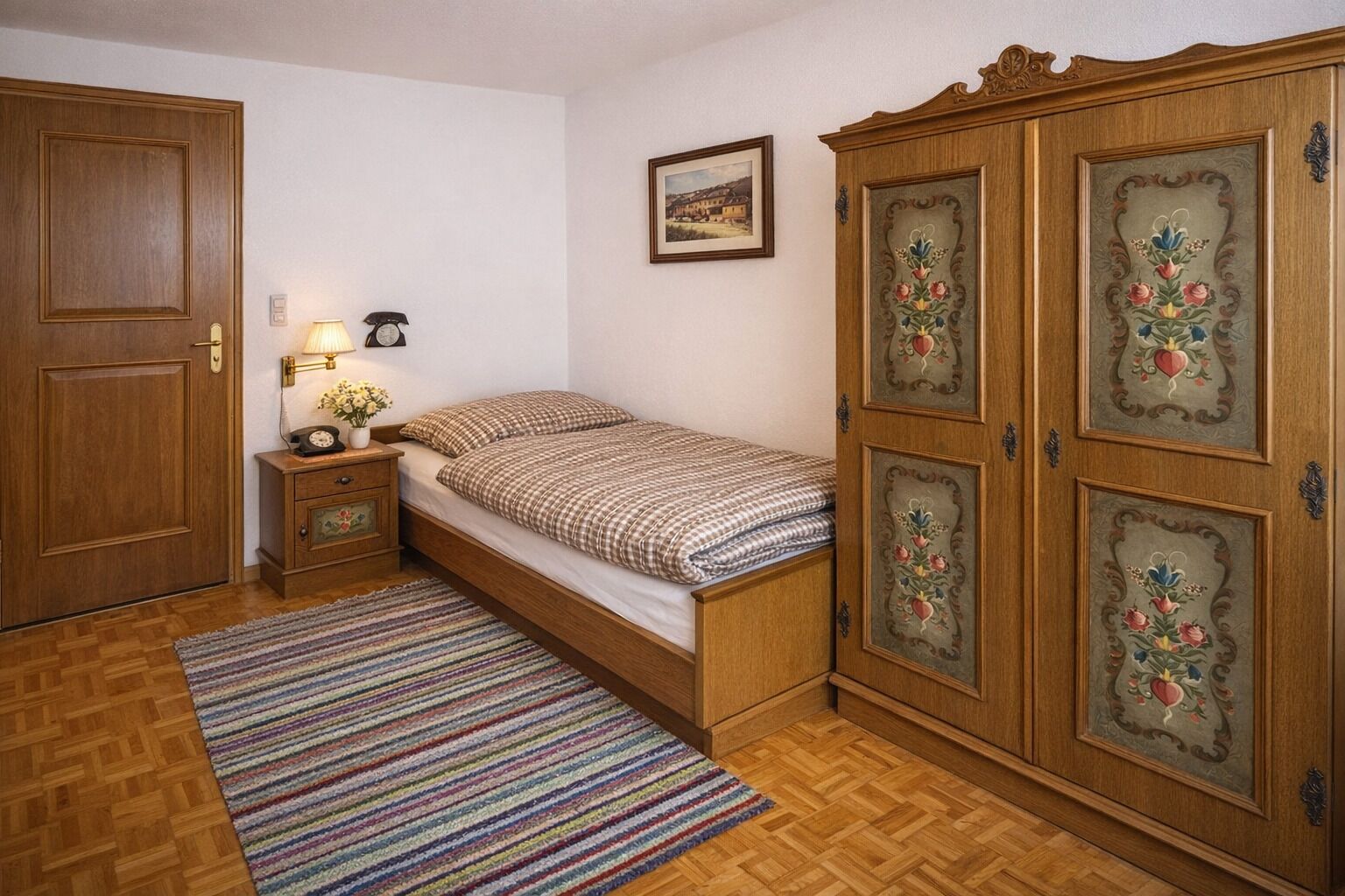Une chambre avec une armoire en bois, un lit avec une literie à carreaux, une table de chevet avec une lampe et un tableau encadré accroché au mur.