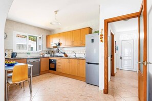 A kitchen with wooden cabinets, a stainless steel refrigerator, a dishwasher, and a dining area with a table and chairs. - Villa La Serena by Villa Plus (Cala en Porter)