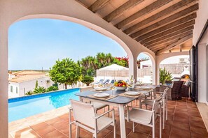 A poolside dining area with a view of the sea, outdoor furniture, and a table set for two. - Villa La Serena by Villa Plus (Cala en Porter)
