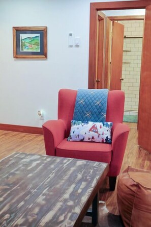 Ein roter Sessel mit einer gesteppten Kissenrolle und einem gemusterten Kissen, ein Holztisch und ein gerahmtes Bild an der Wand.