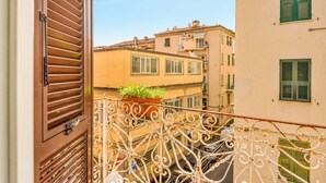 Un balcón con barandilla de hierro forjado, una ventana con persianas y vistas a edificios colindantes.