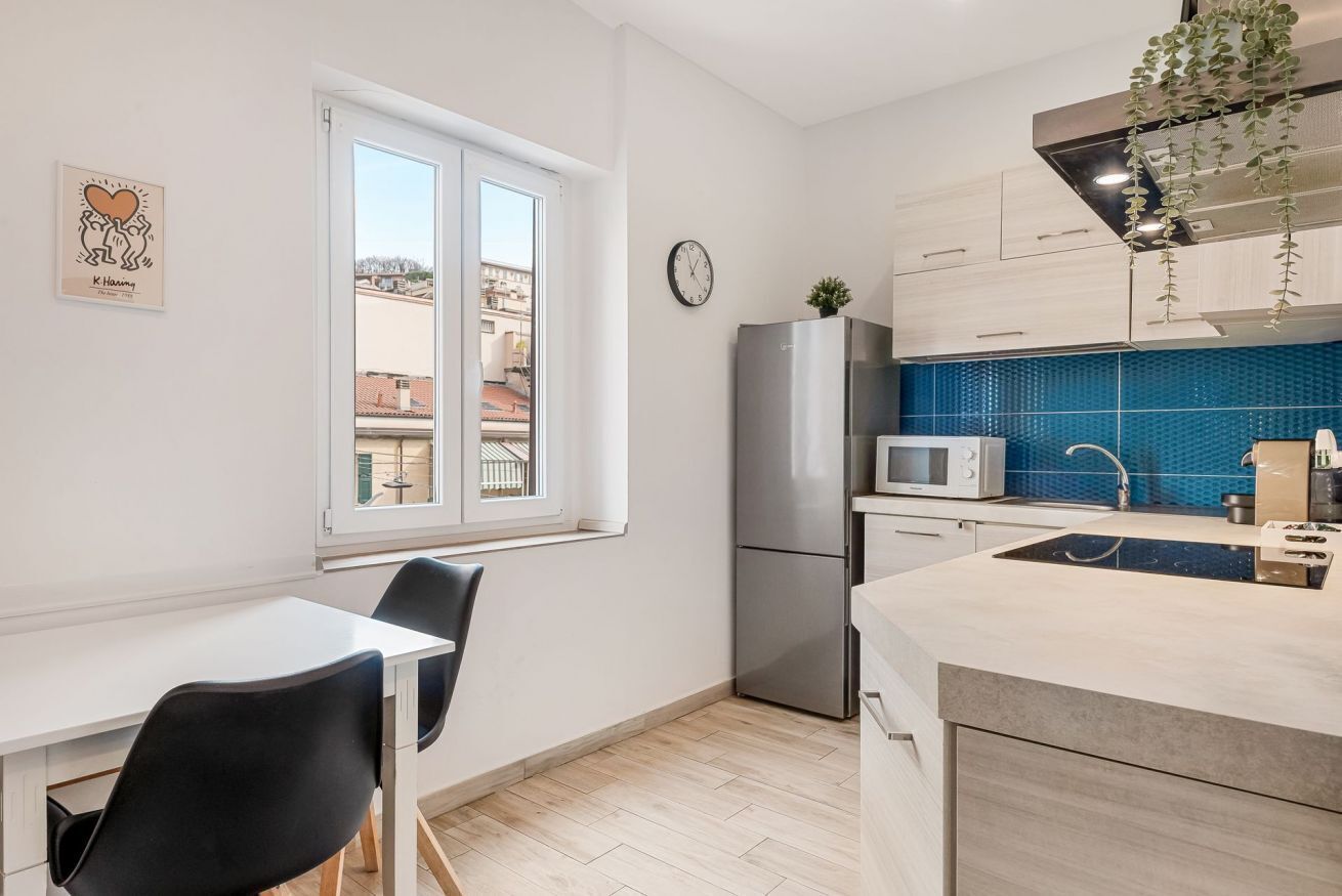 A modern kitchen with a stainless steel refrigerator, a microwave, and a sink with a blue backsplash. There is a window with a view of buildings outside, and a clock on the wall.