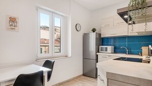 Una cocina moderna con un refrigerador de acero inoxidable, un microondas y un lavabo con un revestimiento azul. Hay una ventana con vista a edificios, y un reloj en la pared.