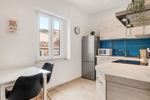 Et moderne køkken med et rustfrit stål køleskab, en mikrobølgeovn og en vask med blå bagplade. Der er et vindue med udsigt til bygninger udenfor, og et ur på væggen.