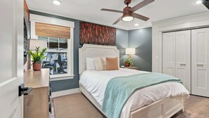 Un dormitorio con cama, ventilador de techo, ventana con persianas, mesita de noche con lámpara y una puerta.