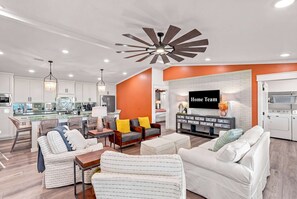 A modern living room with a kitchen in the background, a ceiling fan, and a TV mounted on the wall. - PCB Getaway • Resort Amenities • Golf Cart • Pools (Panama City Beach)