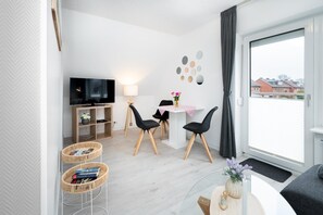 Ein modernes Wohnzimmer mit einem Flachbildfernseher, einem Esstisch mit Stühlen, einer Couch und einem großen Fenster mit Blick auf Häuser.