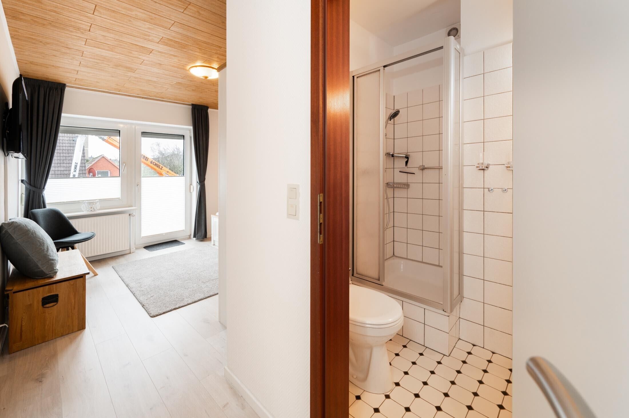 Une pièce avec un plafond en bois, une porte-fenêtre coulissante, une chaise noire, un petit meuble en bois et un plancher blanc.