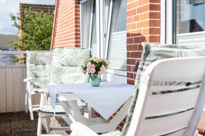 Eine Terrasse mit weißen Stühlen und einem Tisch, eine Vase mit Blumen sowie eine Ziegelwand im Hintergrund.