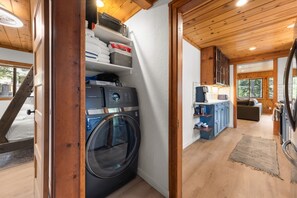 A compact laundry area with a washing machine, shelves with folded towels, and a small kitchenette with a coffee machine. - Homewood Cabin in the Pines — Near the Lake & Homewood Ski Area (Homewood)