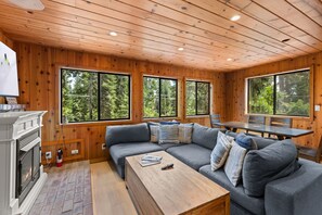 Ein gemütliches Blockhaus-Interieur mit einer Couch, einem Esstisch und großen Fenstern mit Blick auf den Wald.