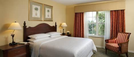 A hotel room with a large bed, two bedside tables with lamps, a chair, a window with curtains, and framed pictures on the wall.