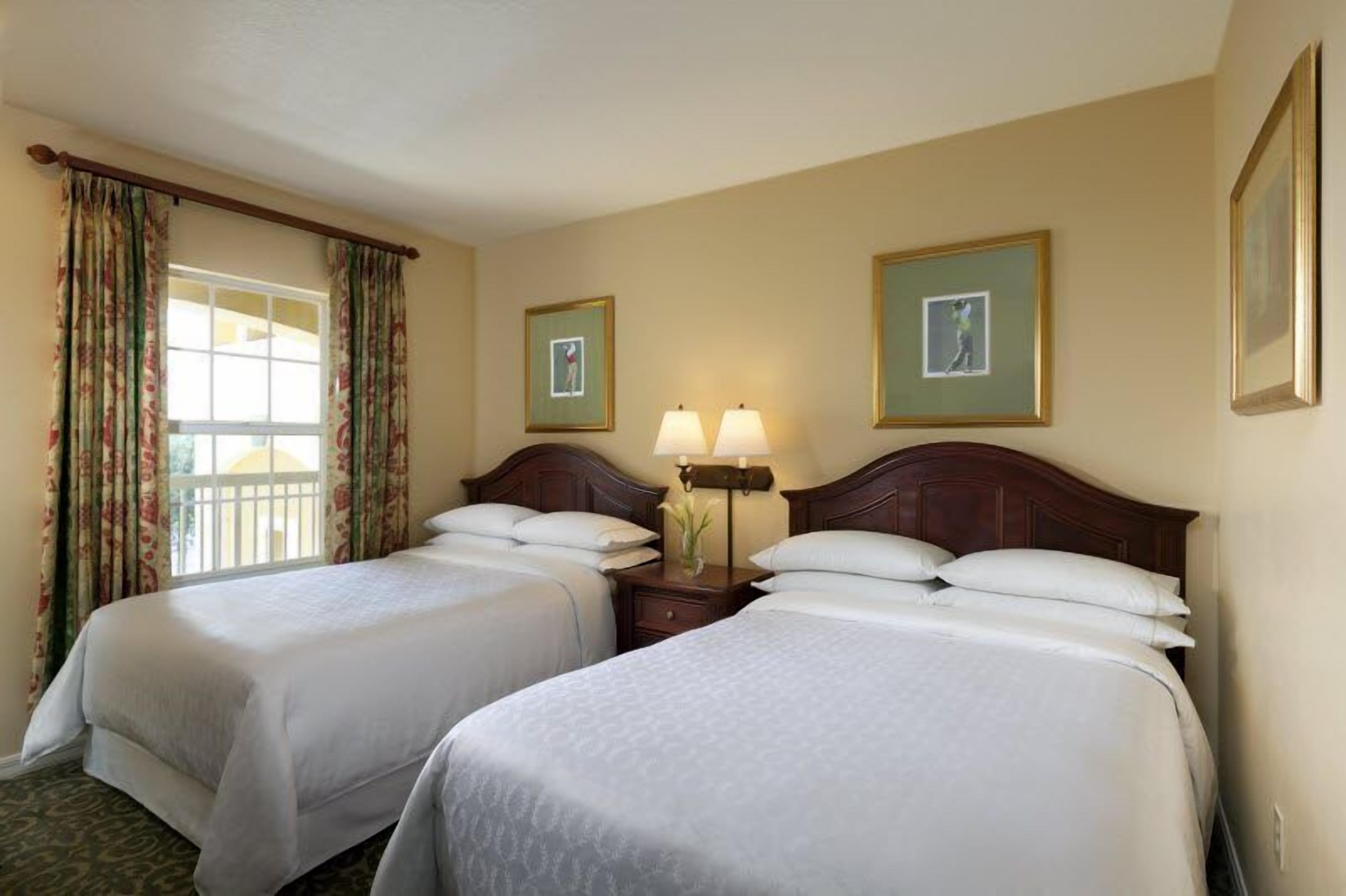 A hotel room with two beds, a nightstand with lamps, and a window with curtains.