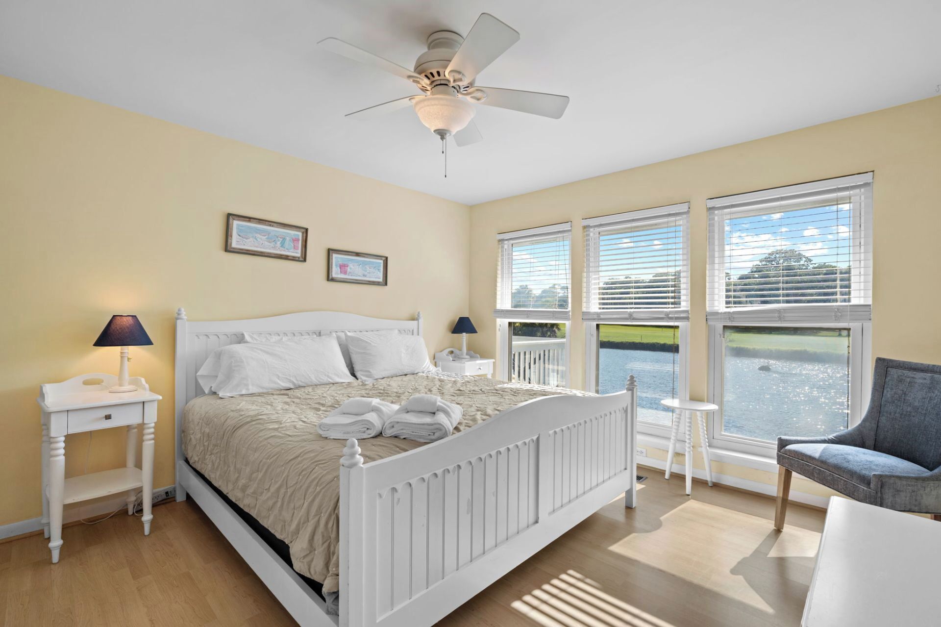 A bedroom with a bed, two bedside tables, a ceiling fan, and a view of a body of water through large windows.