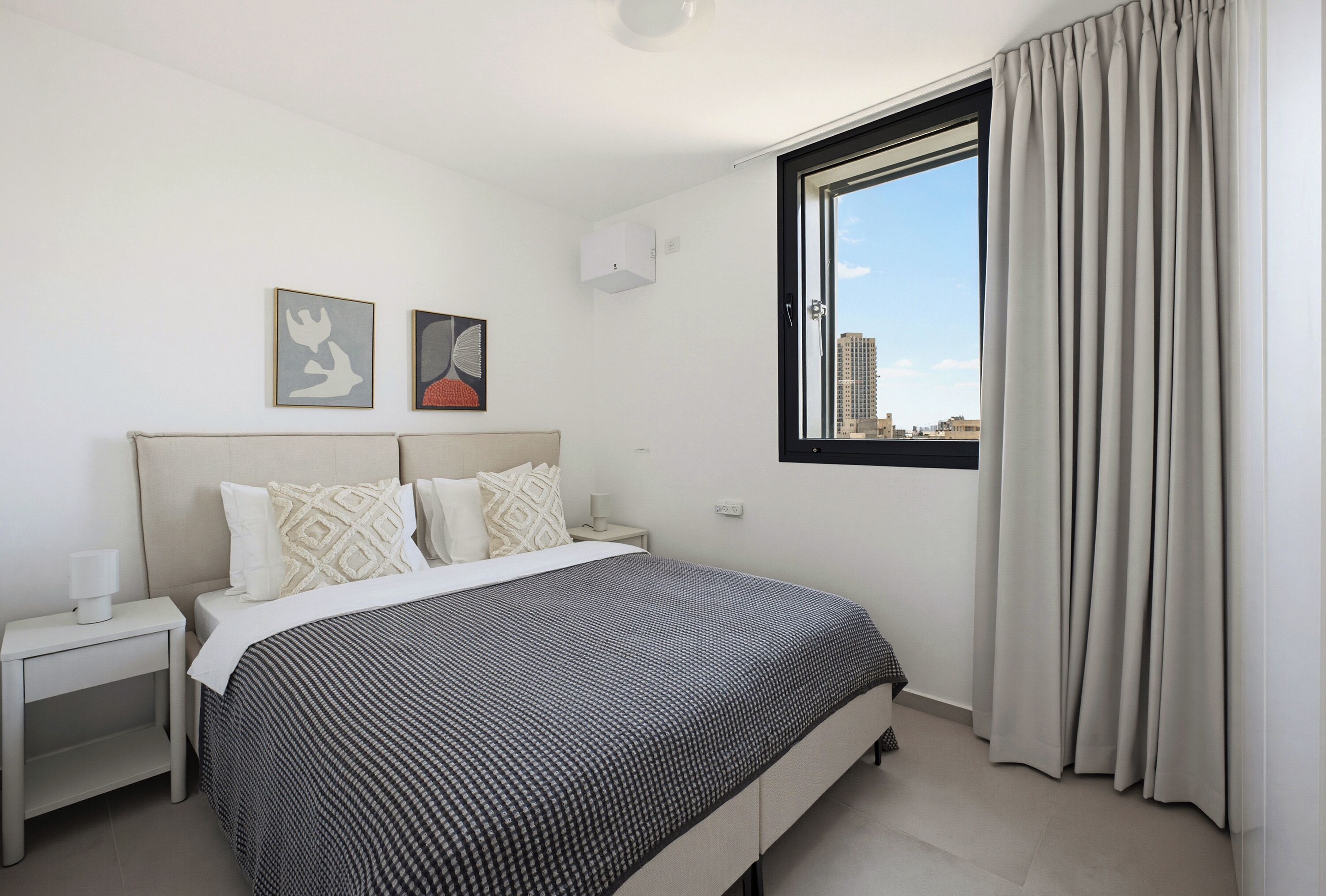 A hotel room with a bed, a nightstand, a window with curtains, and two wall-mounted artworks.