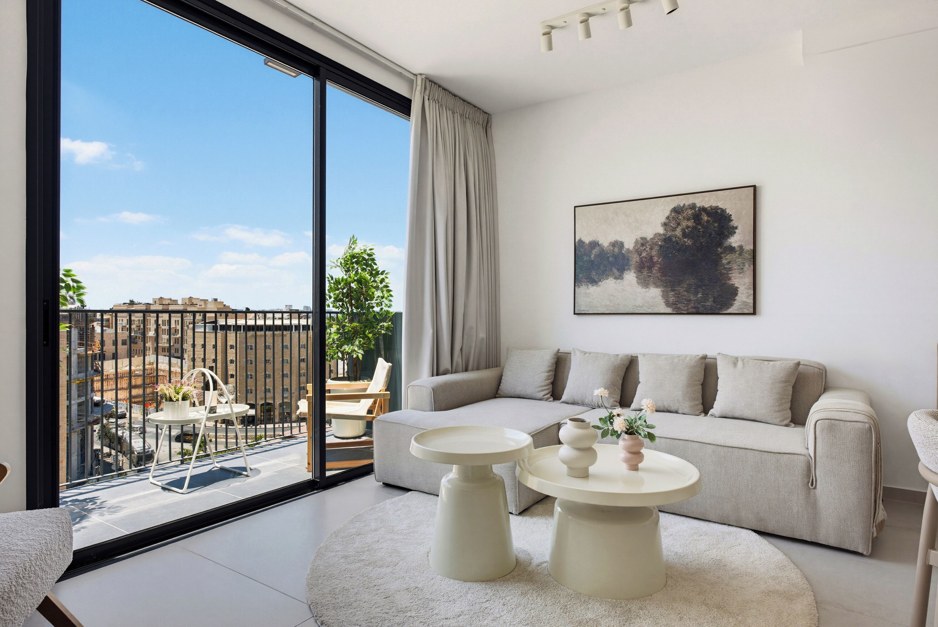 Een moderne woonkamer met een bank, twee bijzettafels, een groot raam met uitzicht op de stad, en een schilderij aan de muur.
