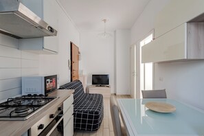 A compact kitchen with a gas stove, a built-in oven, and a microwave. There is a sofa with a black and white checkered cover, a flat-screen TV on a stand, and a dining table with a glass top. - Niguarda Suite (Milano)
