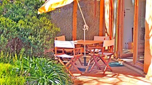 Um pátio externo com mesa e cadeiras de madeira, um guarda-sol amarelo e vista para o verde ao redor.