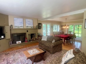 A cozy living room with a fireplace, a dining area with a red tablecloth, and a large window with a view of greenery.