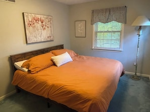 A bedroom with a bed, a window with blinds, a lamp, and wall art.