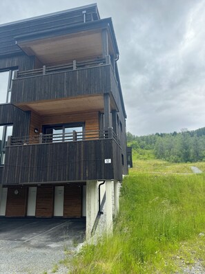 A multi-story building with a balcony on the upper floor, a garage at the base, and a natural landscape in the background. - Beautiful 4-bedroom apartment in peaceful Vestland, perfect for ski and hiking (Vestland)