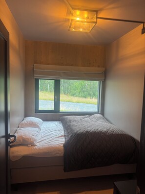 Un lit simple avec une tête de lit, une fenêtre donnant sur un paysage verdoyant et un luminaire au plafond.