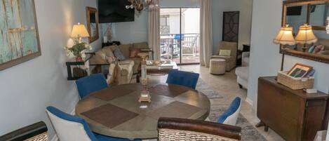 A spacious living area with a round dining table, comfortable seating, a television, and a balcony view.