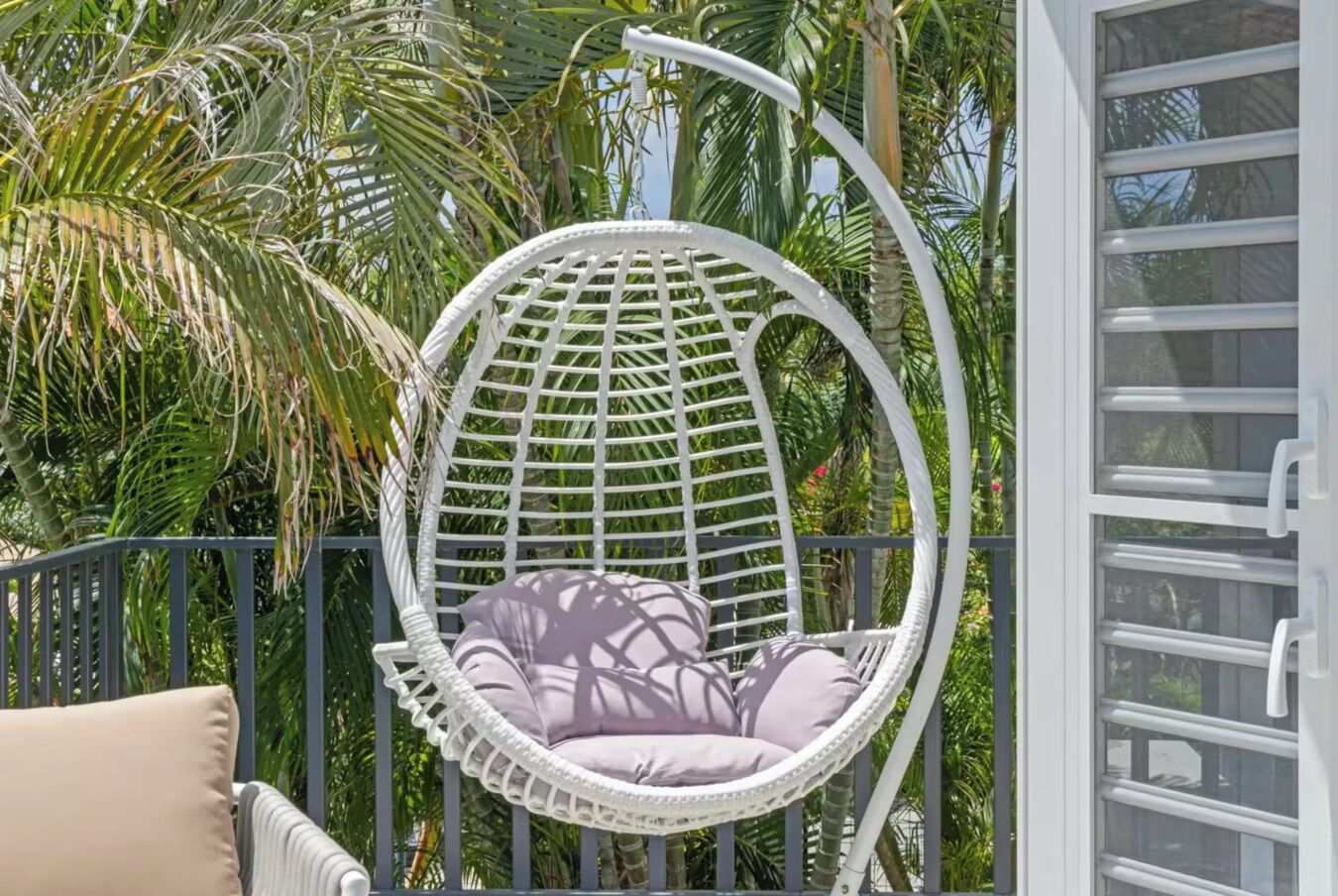 A white wicker swing chair with cushions, positioned near a window with a metal railing.