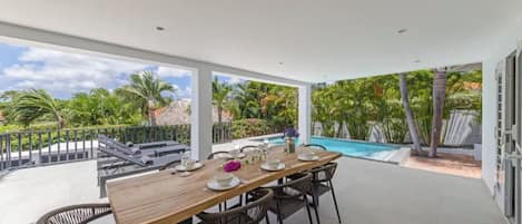 An outdoor dining area with a pool, surrounded by greenery and a clear sky.