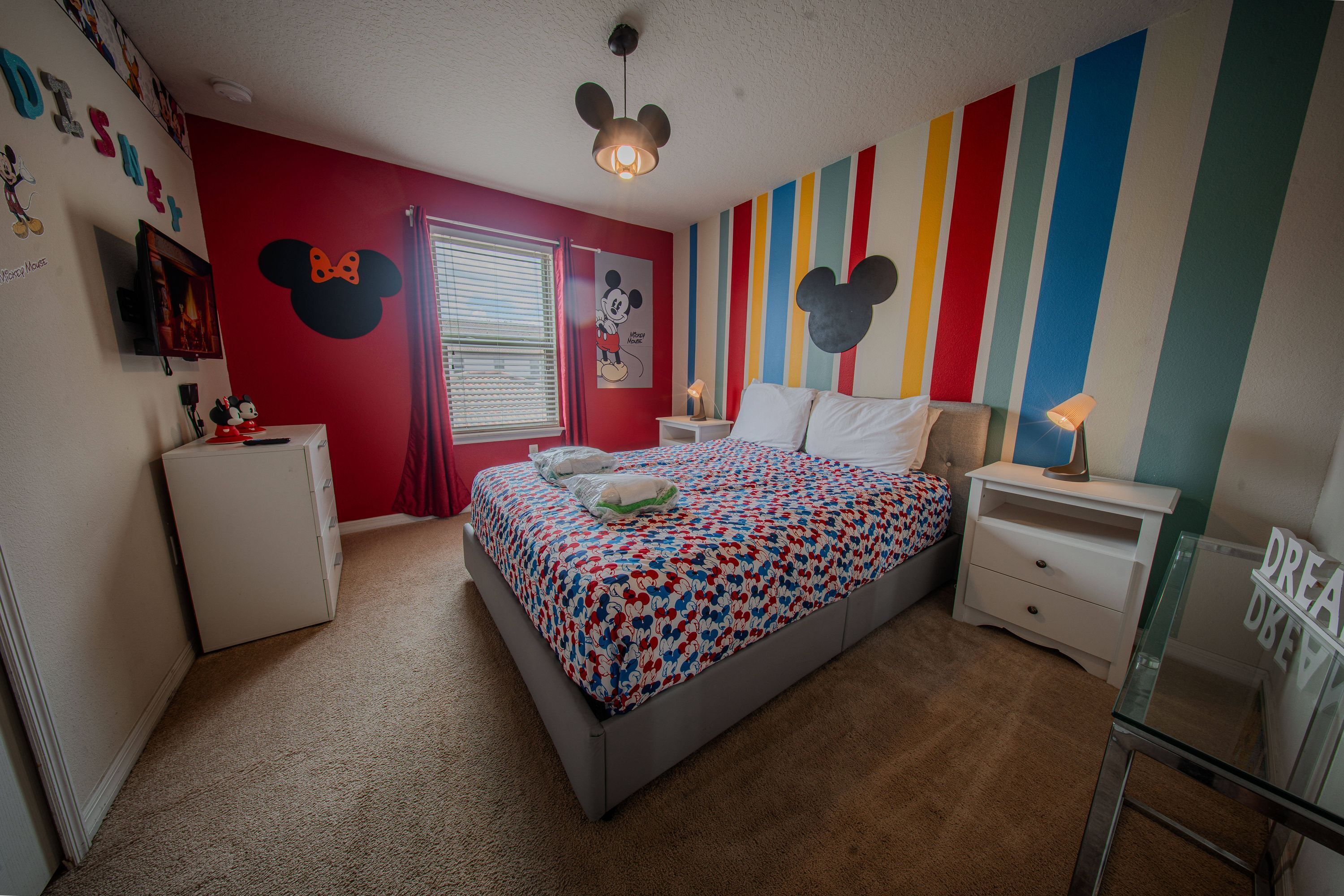 Un dormitorio con cama, mesita de noche, cómoda y una pared con temática de Mickey Mouse, además de una ventana con persianas.