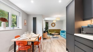 A modern kitchen with dark cabinetry, a dining area with a wooden table and orange chairs, and a living room with a green sofa and blue armchairs. - Woodland View 2 By My Getaways (Brighton)