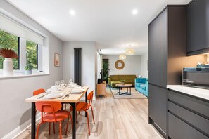 A modern kitchen with dark cabinetry, a dining area with a wooden table and orange chairs, and a living room with a green sofa and blue armchairs. - Woodland View 2 By My Getaways (Brighton)