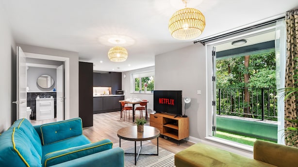 A modern living room with a sofa, a coffee table, and a television. There is a dining area with a table and chairs. The room has a large window with a view of greenery outside. - Woodland View 2 By My Getaways (Brighton)