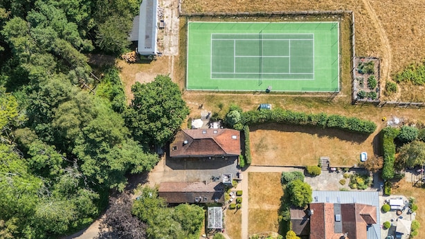 Vista aérea de una zona residencial con cancha de tenis, una casa de techo inclinado y vegetación circundante.