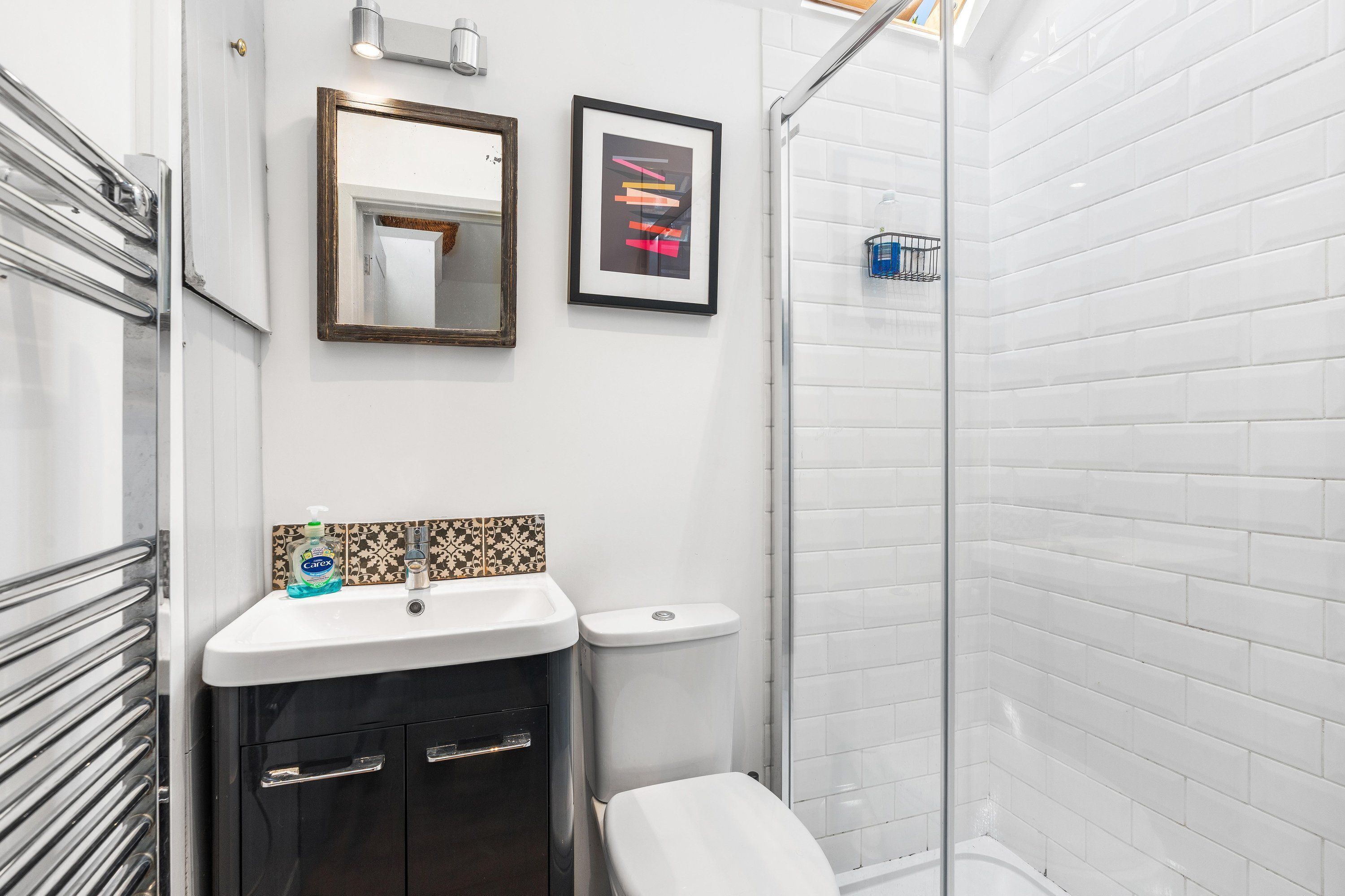 A bathroom with a white sink, a toilet, a shower with white tiles, a mirror, and a framed picture on the wall.