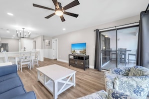 Ein modernes Wohnzimmer mit Deckenventilator, ein Essbereich mit weißem Tisch und Stühlen, eine Küche mit weißen Schränken und ein Balkon mit Glastür.